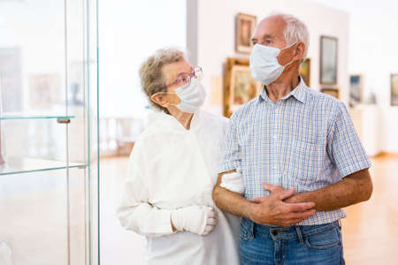 elderly European couple in mask protecting against covid examines paintings on display in hall of art museumの写真素材