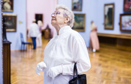 elderly European woman examines paintings in an exhibition in hall of an art museumの写真素材