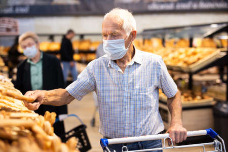 older european man wearing mask and gloves with covid protection chooses buns and bread in supermarket bakeryの写真素材