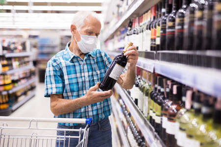 older european man wearing mask with covid protection chooses bottle of alcohol in supermarketの写真素材