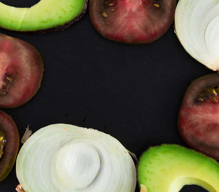 frame of ripe chopped vegetables black tomatoes, avocado and onions against black stone board