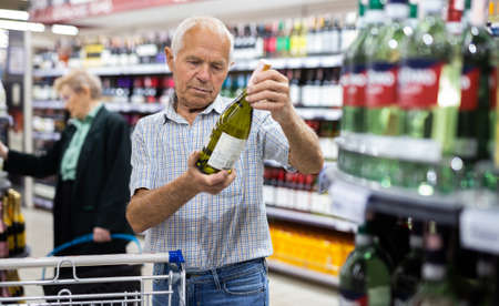 mature european man chooses bottle of white dry wine in alcohol section of supermarketの写真素材
