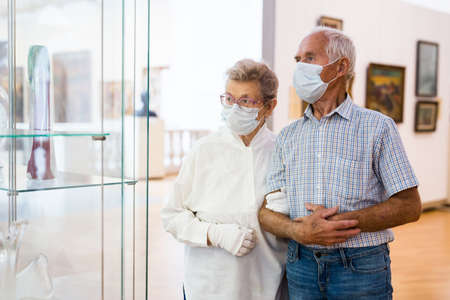 elderly European couple in mask protecting against covid examines paintings on display in hall of...
