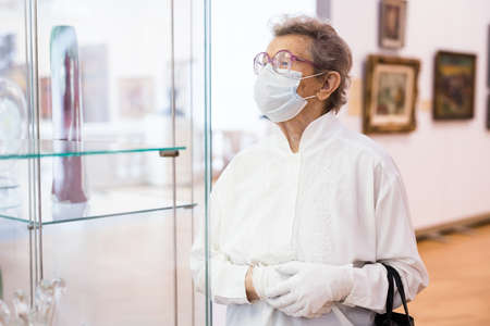 mature woman in mask protecting against covid examines paintings on display in hall of art museum