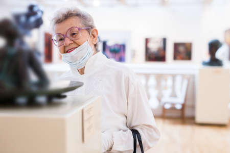 mature woman in mask protecting against covid examines paintings on display in hall of art museum
