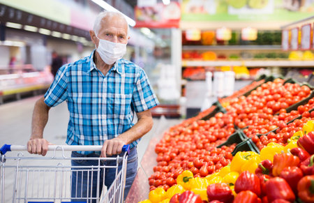 mature caucasian man in mask with covid protection choosing tomatoes in vegetable section of supermarketの写真素材