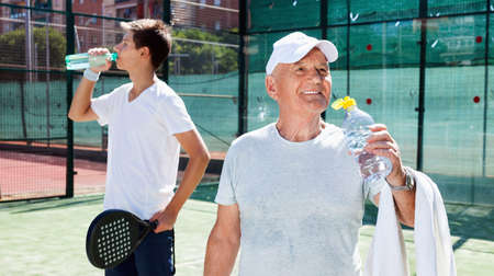 Glad positive smiling mature man and young man drink water on padel courtの写真素材