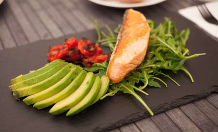 Fried salmon fillet with arugula, avocado and cherry tomatoes on black stone board