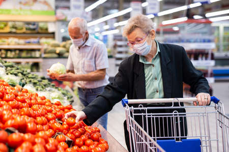 mature caucasian woman in mask and gloves with picks tomatoes in vegetable section of supermarketの写真素材