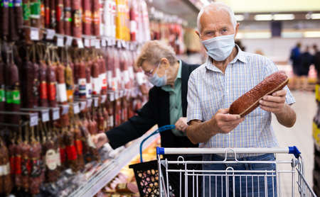 older european man in mask and gloves with covid protection picks up salami in meat section of supermarketの写真素材