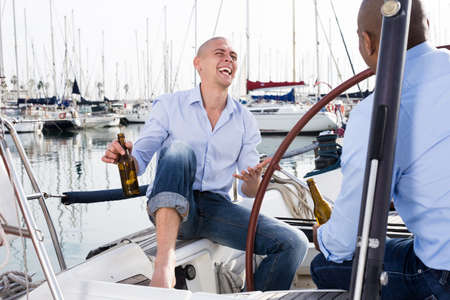 Two men in blue shirts and jeans with bare legs drinking beer and having fun on a yacht in the portの写真素材