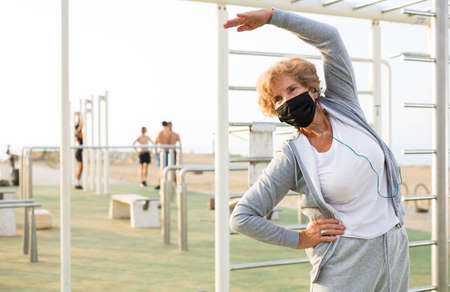 Old lady in face mask doing fitness exercises in outdoor workout station.