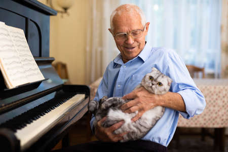 Elderly european man sitting beside piano with cat in hands.の写真素材