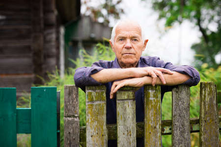 Old man leaning on fence near his country house and looking in camera.