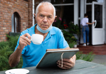 Senior man in mask on chin sitting at table and drinking tea in outdoor cafe. He's holding white cup...