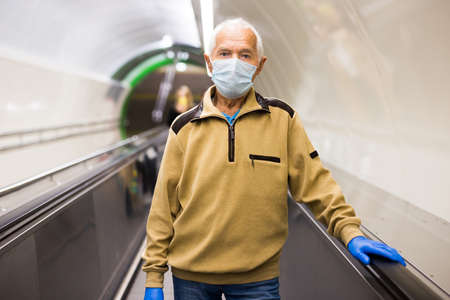 Senior man in face mask and protective gloves standing on moving walkway in metro station.の写真素材