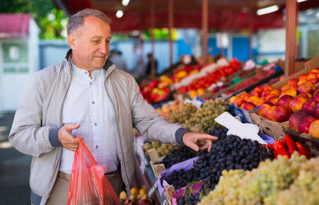 European man standing in fruit shop beside counter and choosing grape.の写真素材