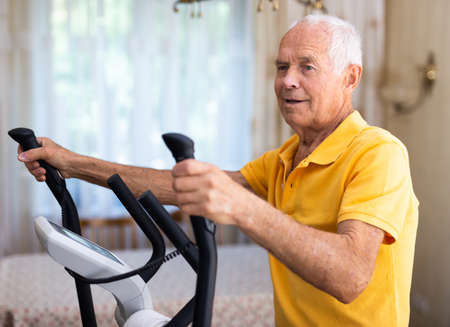 Sportive old man using elliptic trainer to warming up at home.の写真素材