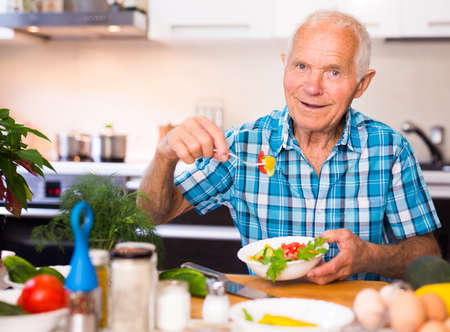 european elderly man cuts vegetables for salad at the table in the kitchenの写真素材