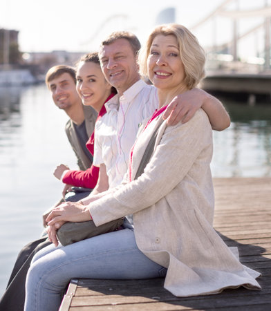 family of tourists sitting on embankment dangling legs above the water in port in European cityの写真素材
