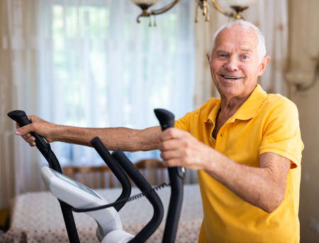 Positive senior man works out on an elliptical machine at homeの写真素材