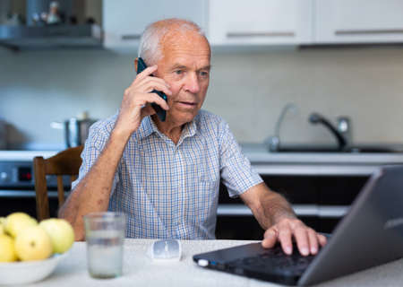 Mature man buys stock online by mobile phone using laptop and internet while sitting in kitchen of his homeの写真素材