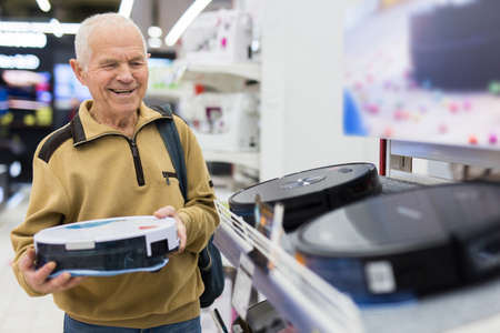 elderly gray-haired man pensioner looking robot at counter in showroom of electrical appliance hypermarket departmentの写真素材