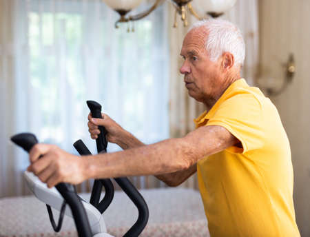 Elderly man exercising on elliptic trainer in living roomの写真素材