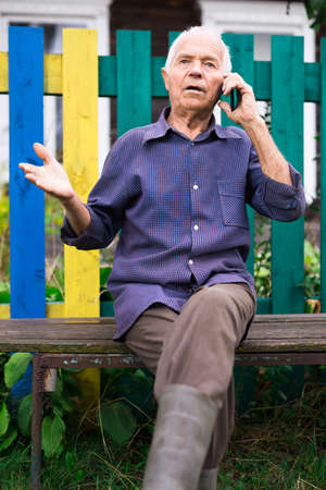 Elderly man sits on bench near his country house and talks on mobile phoneの写真素材