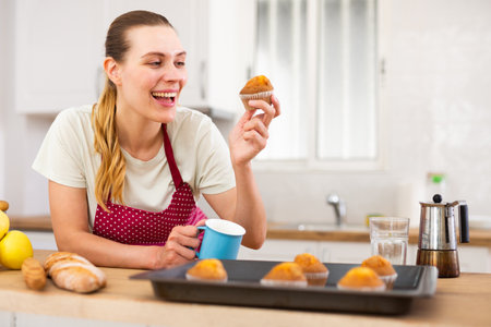 Woman in apron tasting freshly baked sweet cupcakes in kitchenの写真素材