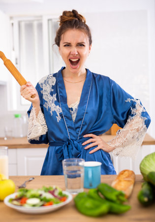 Angry woman screaming and waving rolling pin in home kitchenの写真素材