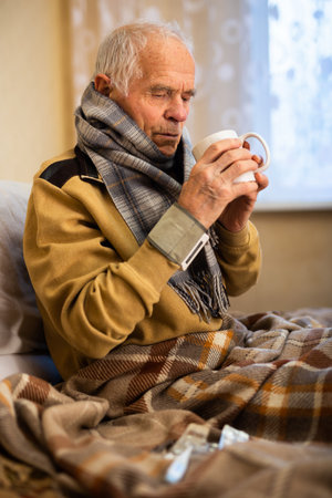 old man wrapped in blanket has cold and drinks hot infusionの写真素材