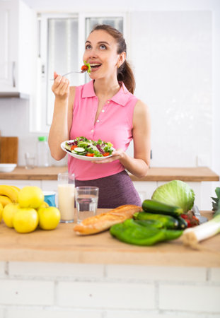 Slim young woman eating vegetable salad in home kitchenの写真素材