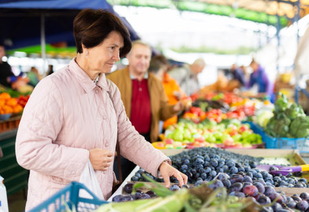 Positive mature woman in casual clothes picking fresh plums during shopping at grocery marketの写真素材