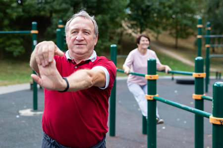 Staying after retirement. Happy joyful mature retired sportsman wearing sportswear doing side stretching exercises with arm over his head, exercising outside in city parkの写真素材