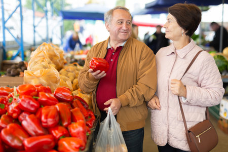 Positive senior couple in casual wear picking bell peppers during shopping at crowded bazaarの写真素材