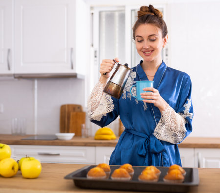 Pretty woman in dressing gown drinking coffee and eating muffins in kitchen during breakfastの写真素材