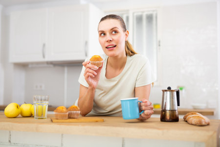 Happy young woman enjoying cupcakes with coffee in kitchenの写真素材