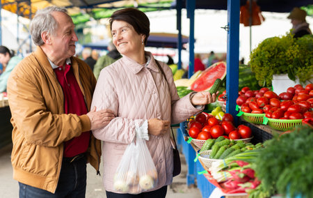 Positive mature man and woman choosing organic cucumbers near vegetable stand at local grocery marketの写真素材