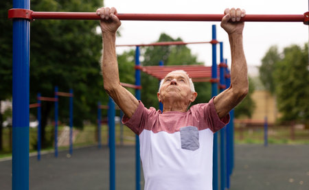 Old man pulls himself up on horizontal bar on sports ground. Outdoor sports in old ageの写真素材