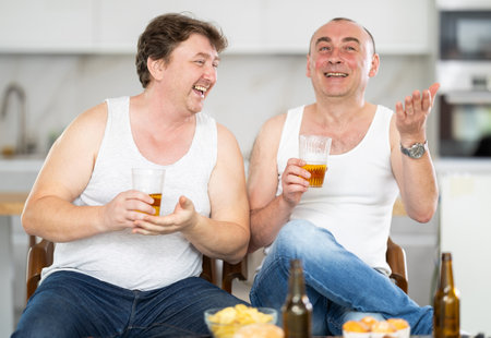 Two adult male friends drinking beer and eating chips at home. They are happy and enjoying each other's company.の写真素材