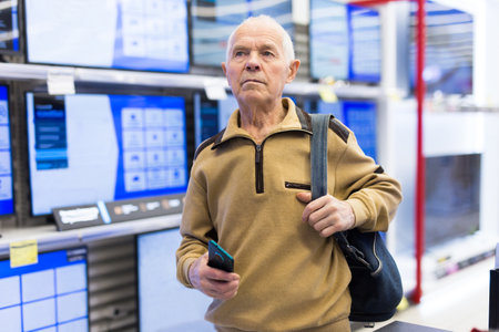 Elderly grayhaired man pensioner looking counter with modern digital televisors in showroom of digital goods storeの写真素材