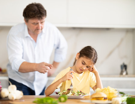 Father forcing little daughter to eat vegetable salad in kitchenの写真素材