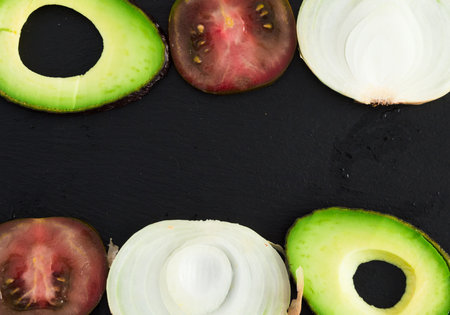 Frame of chopped vegetables black tomatoes, avocado and onions against black stone boardの写真素材