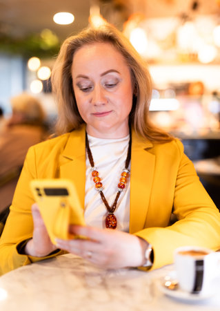 Woman sits at table in restaurant, drinks coffee and writes a message on phone.の写真素材