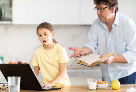Father invites his little daughter to read an interesting book together. Daughter refuses and continues to play games on laptopの写真素材