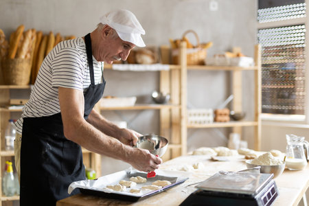 Mature baker brushing uncooked dough pieces on baking sheet with oil before baking in ovenの写真素材