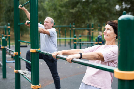 Elderly man and woman doing exercises on horizontal bars at outdoor sports groundの写真素材