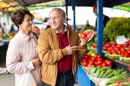 Positive senior couple in casual wear picking tomatoes during shopping at crowded bazaarの写真素材