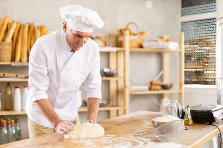 Elderly male baker preparing raw yeast dough on kitchen table in bakeryの写真素材
